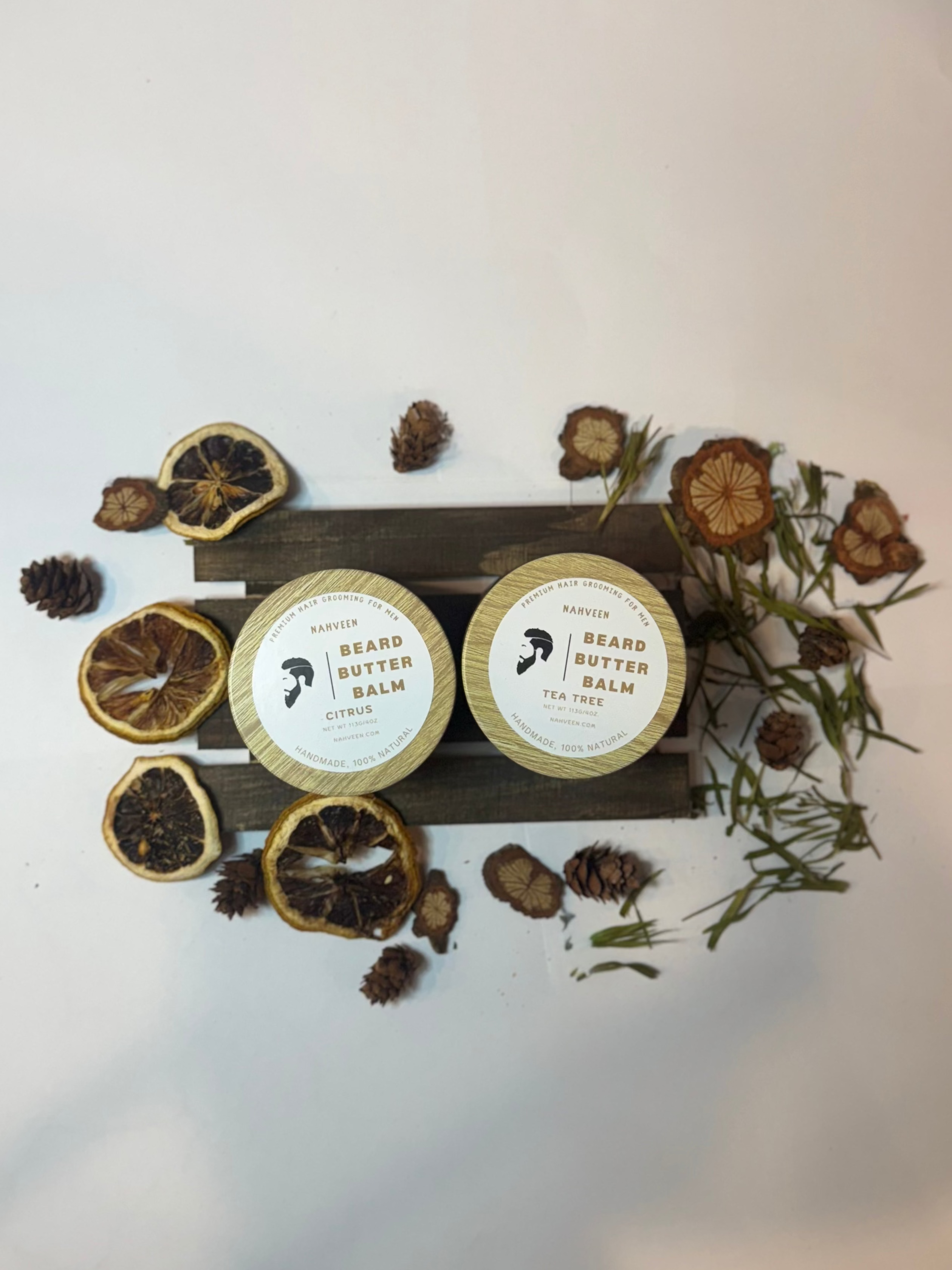 Two round containers labeled 'Beard Balm' on a wooden stand with dried herbs and leaves on a white background